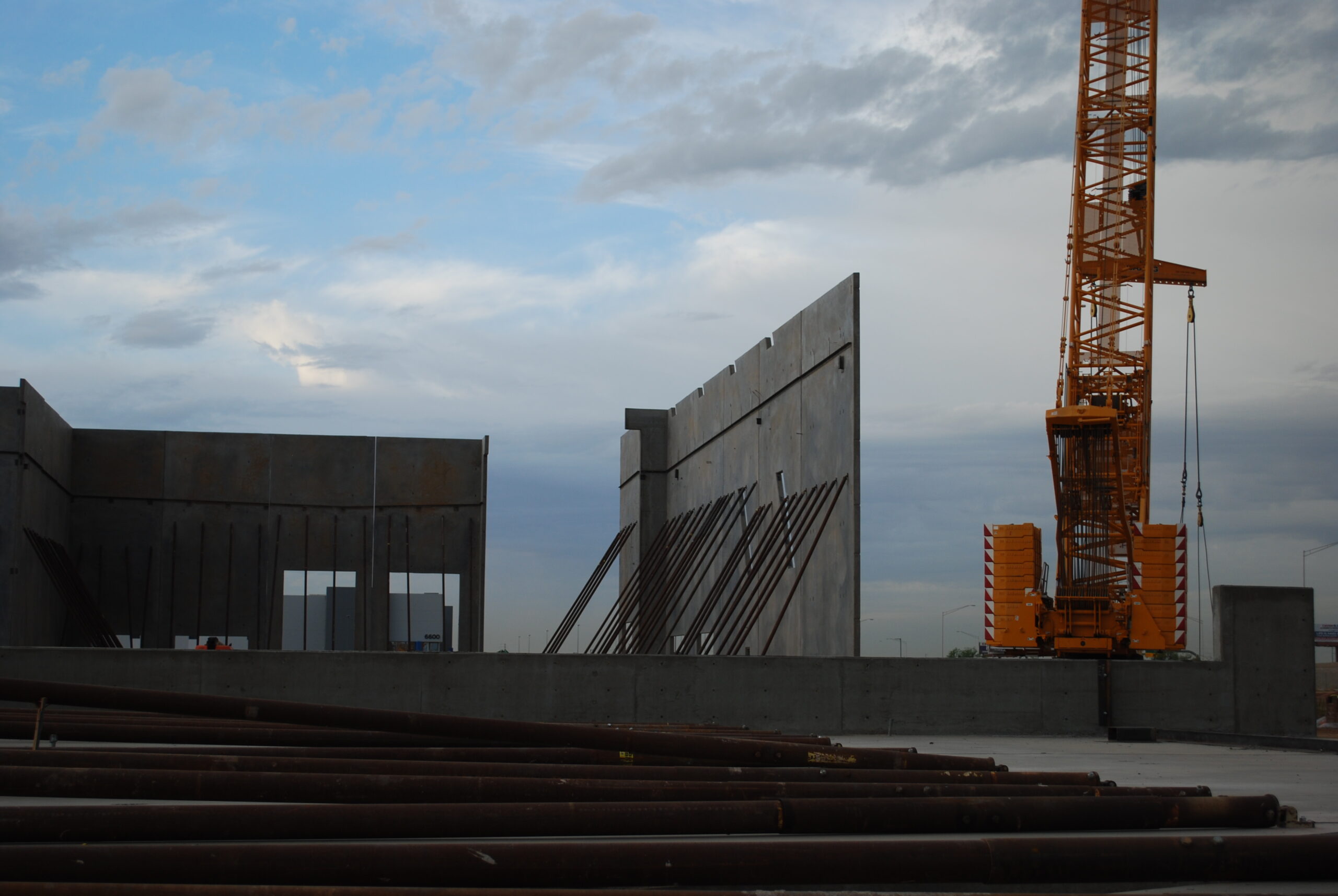 A construction site with concrete walls being installed, steel rods on the ground, and a large yellow crane under a partly cloudy sky.