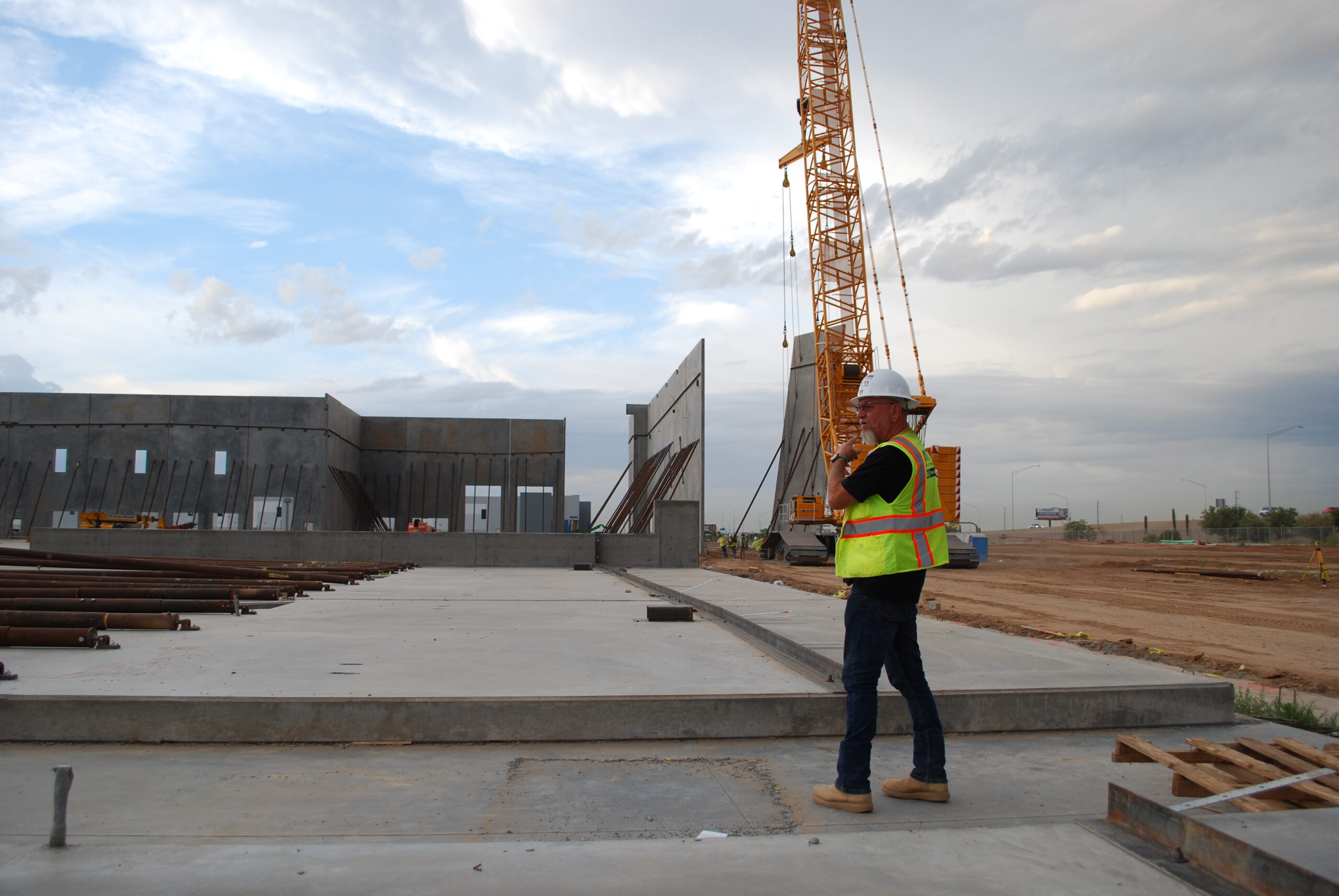 A construction worker in a safety vest and helmet stands on a concrete slab near a large crane at an active construction site.